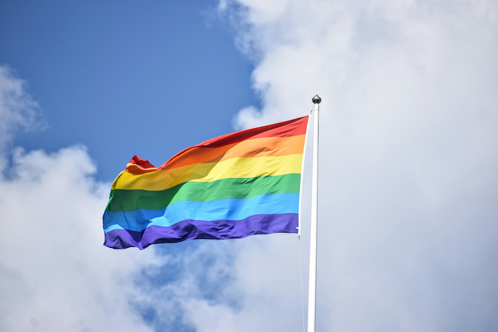 A rainbow Pride flag lifting in the wind against a bright blue sky with soft clouds.
